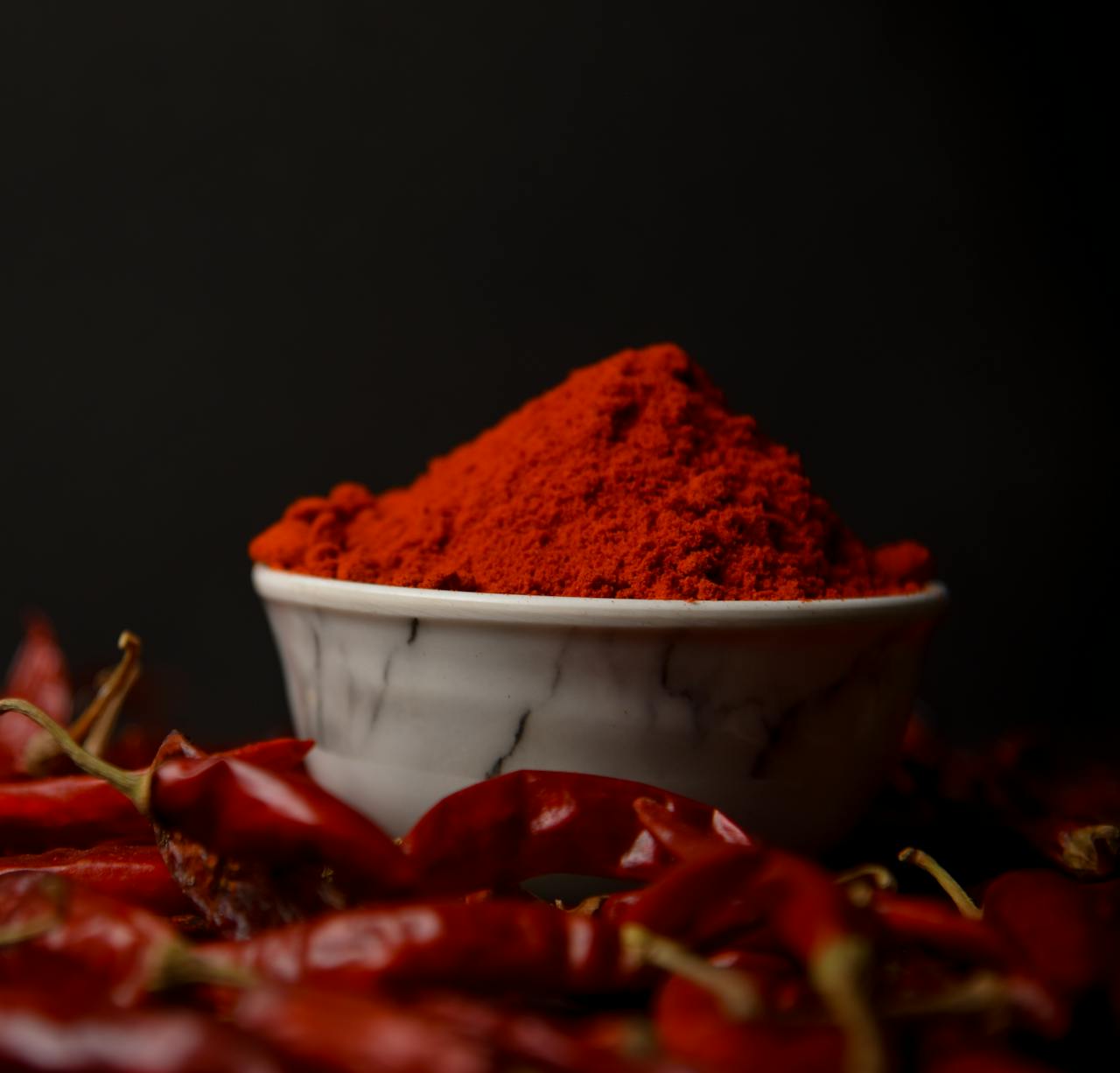 A bowl of vibrant red chili powder surrounded by whole dried chilies on a dark background.