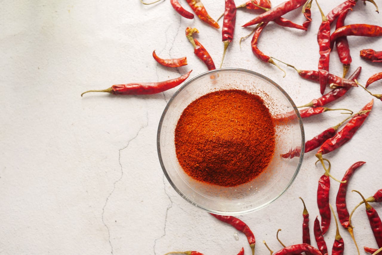 Bowl of vibrant chili powder surrounded by red chili peppers on a textured white background.